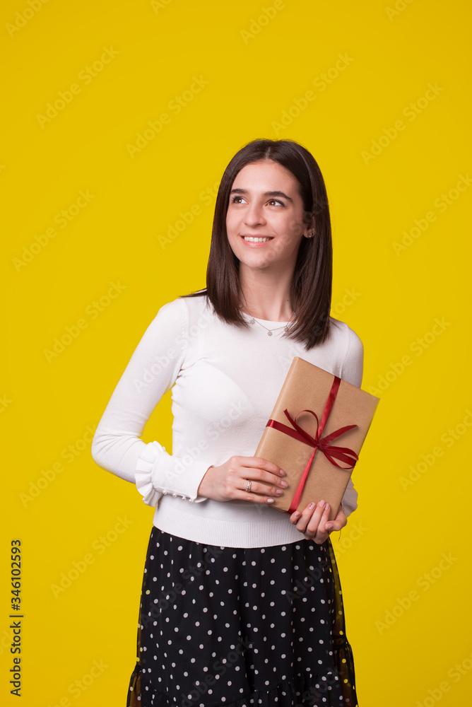 Fototapeta premium Young lovely smiling woman is holding a wrapped gift looking far on yellow background.