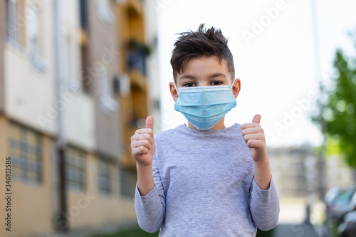 7 years old boy in medical face mask shows thumbs up. Boy having fun during the quarantine. Stop novel coronavirus or stop covid-19 concept
