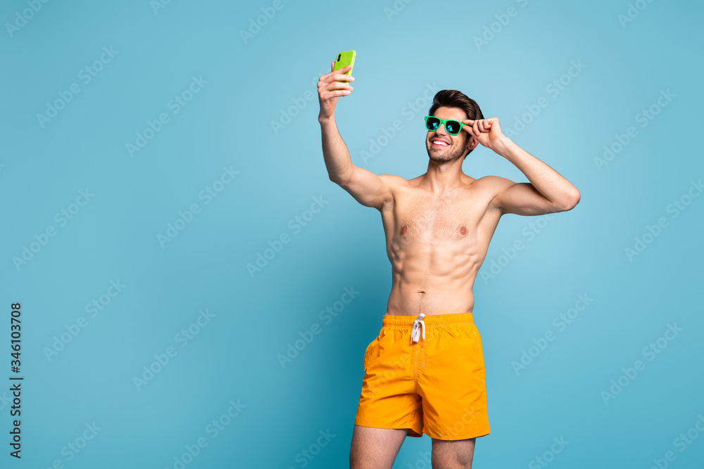 Photo of cheerful positive handsome man adjusting his eye glasses taking selfie recording video with telephone in yellow shorts isolated pastel blue color background