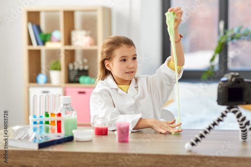 science, childhood and blogging concept - happy smiling little girl blogger with slime and camera on tripod recording video blog at home laboratory