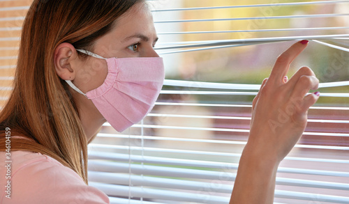 Young woman in pink home made cotton virus face mask, looking through window blinds outside. Quarantine or stay at home during coronavirus covid 19 outbreak