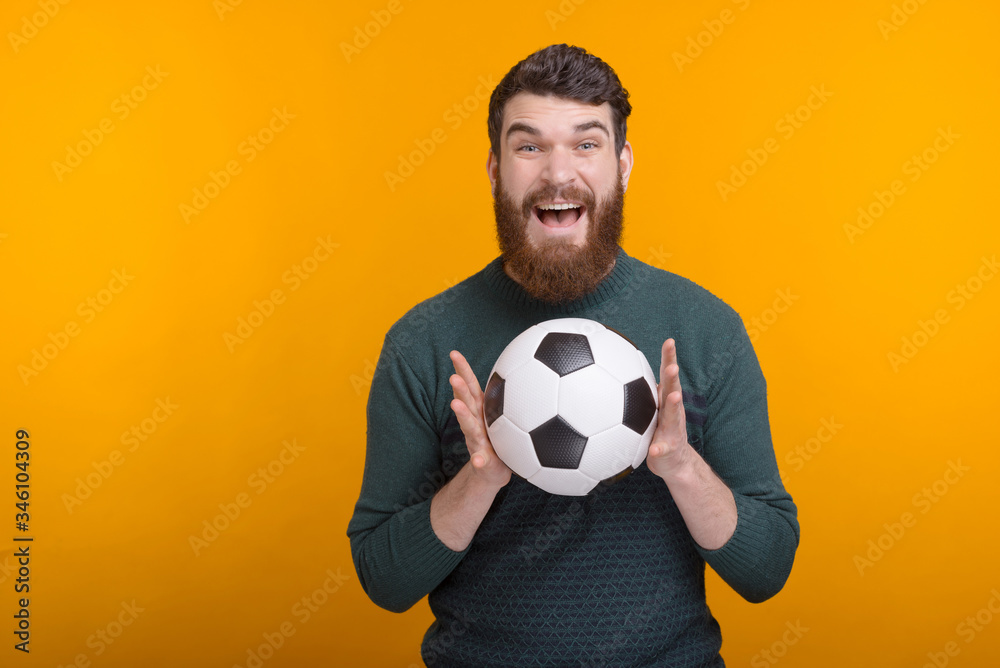 Surprised bearded man is holding with both hands a soccer or football ball on yellow background. Surprised bearded man is holding with both hands a soccer or football ball on yellow background.