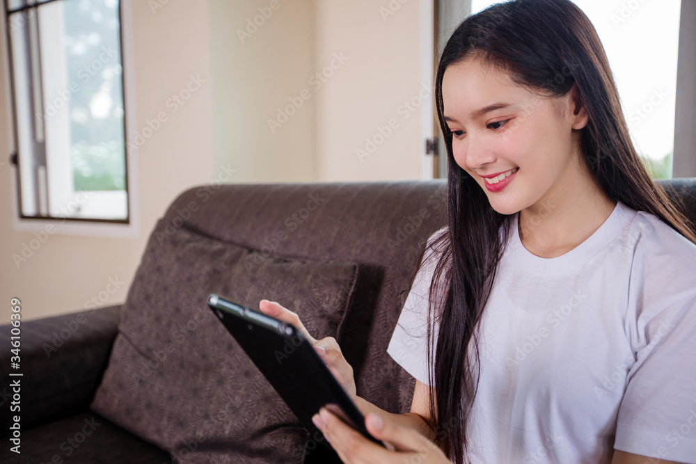 Asian young woman happy cheerful cute beautiful is playing digital pad tablet computer and sitting on the sofa at home, smiling lady using app reading electronic book relax in living room Asian young woman happy cheerful cute beautiful is playing digital pad tablet computer and sitting on the sofa at home, smiling lady using app reading electronic book relax in living room