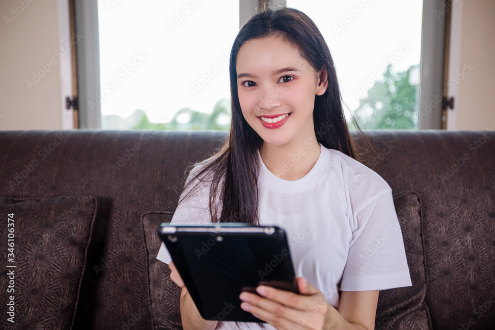 Asian young woman happy cheerful cute beautiful is playing digital pad tablet computer and sitting on the sofa at home, smiling lady using app reading electronic book relax in living room Asian young woman happy cheerful cute beautiful is playing digital pad tablet computer and sitting on the sofa at home, smiling lady using app reading electronic book relax in living room