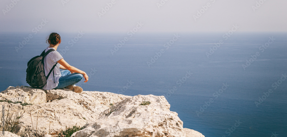 A girl traveler sits on a rock and admires the blue boundless sea, freedom, travel, unity with nature, banner panorama format