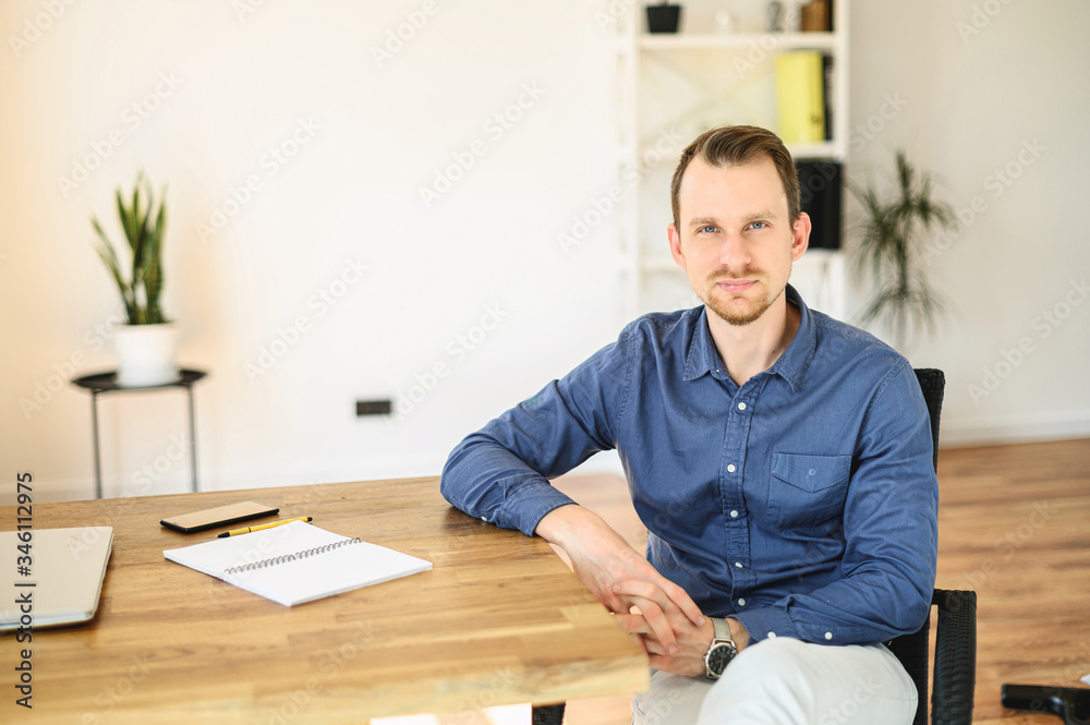 Office work. Portrait of young confident businessman in smart casual wear, a guy sits at the table and looks at camera with a smile