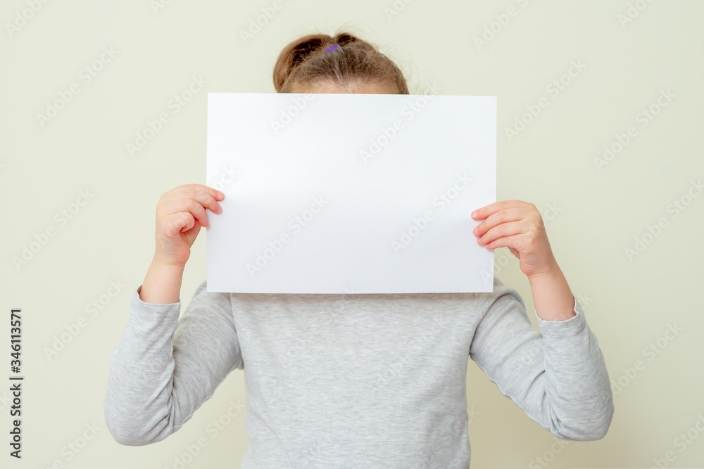 Clean sheet of a paper in hands of child covering her face on yellow background. Mockup sheet of paper in child's hands. Clean sheet of a paper in hands of child covering her face on yellow background. Mockup sheet of paper in child's hands.