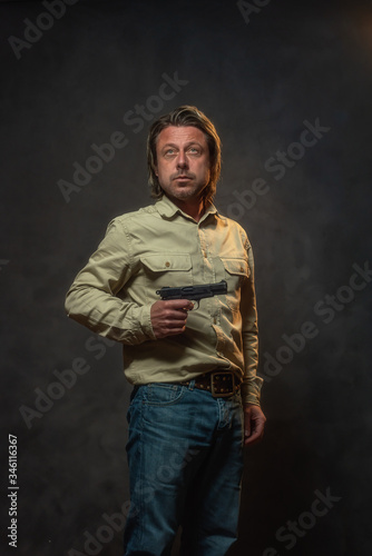 Man with pistol in light brown shirt and jeans in front of grey wall. Low key studio shot.