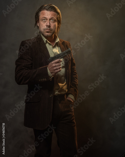 Fashionable middle aged man with handgun in dark brown suit in front of grey wall. Low key studio shot.