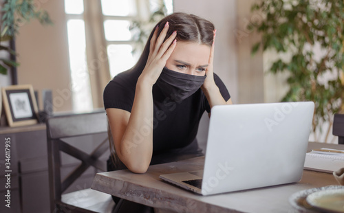 A girl in a medical black mask works at a computer and surf the internet. A young woman feels annoyed and tired.
