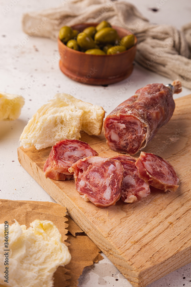 Sliced salami, bread, olives and cutting board on a white background, seen from the front