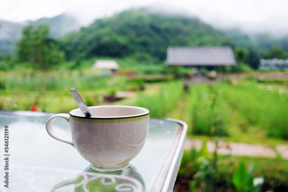 A cup of hot coffee on glass table in the tropical garden with valley mountain, farm and rainforest with fog cover on hill view in rainy season at Thailand