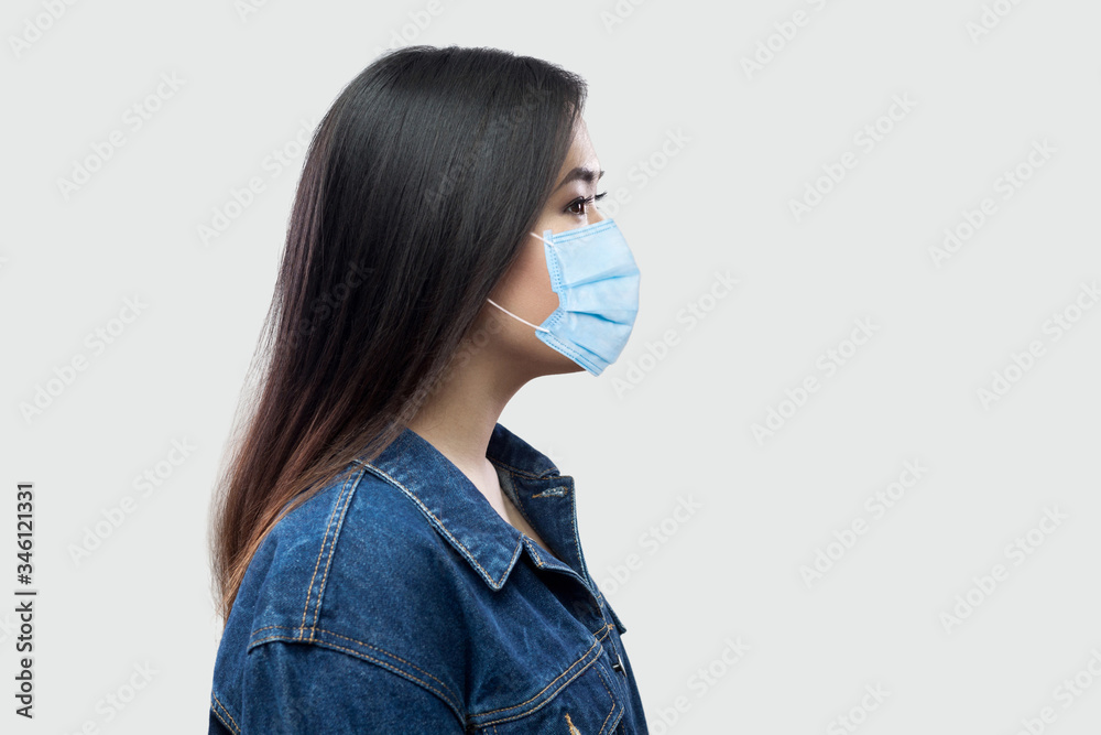 Profile side view portrait of calm serious beautiful brunette asian young woman with surgical medical mask in blue jacket standing and looking. indoor studio shot, isolated on light grey background. Profile side view portrait of calm serious beautiful brunette asian young woman with surgical medical mask in blue jacket standing and looking. indoor studio shot, isolated on light grey background.