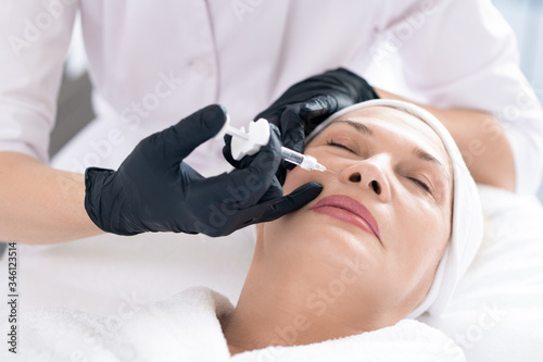 Close-up of unrecognizable cosmetologist using syringe while injecting filler in women's face to rejuvenate her skin