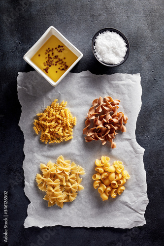 Variety of raw pasta with olive oil and salt on baking paper on black background photo