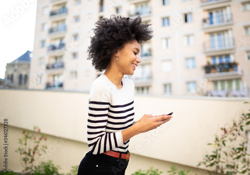 Side of smiling young black woman walking with mobile phone outside