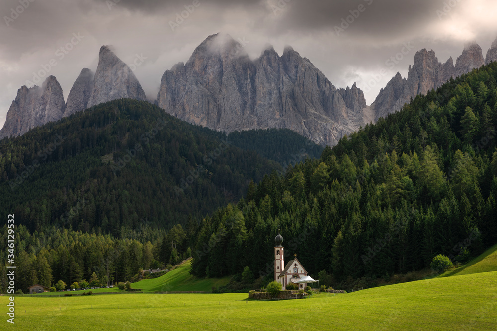 Mountain panorama with church St. Johann in St. Magdalena, sun on green meadow, forest and mountains in the background, heavy clouds in the sky, Val Di Funes, Dolomite Alps, South Tyrol Italy.