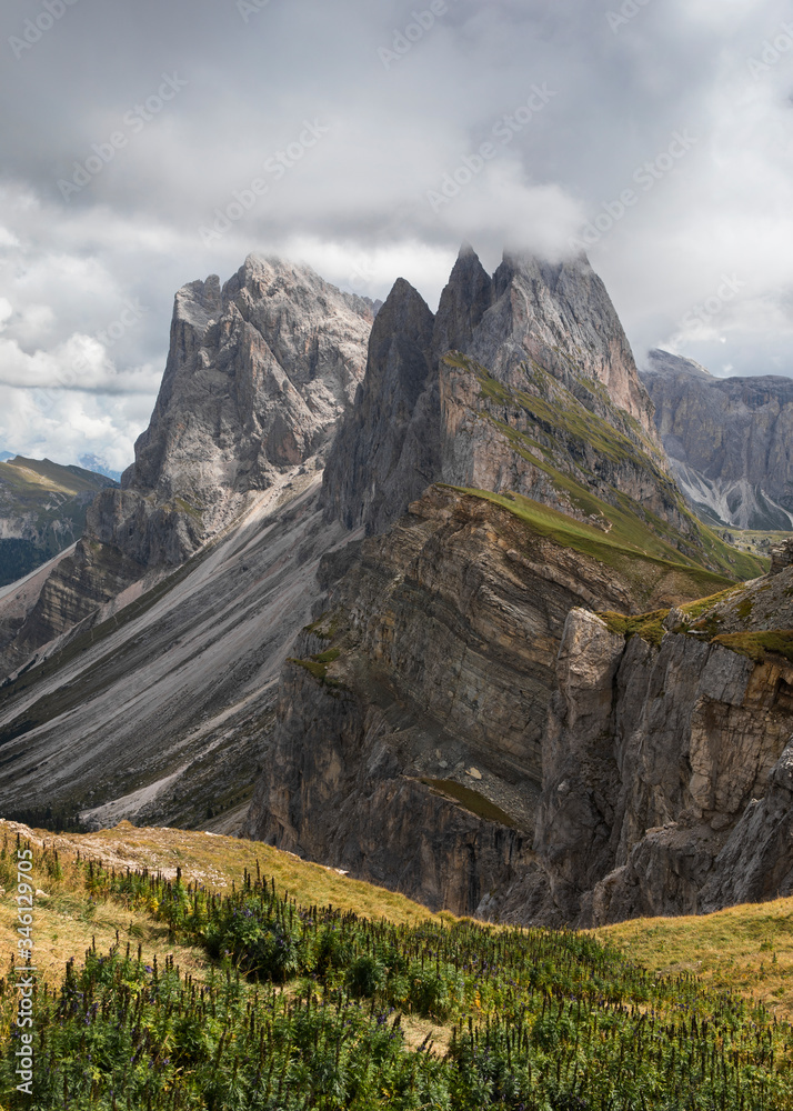 Naklejka premium Dramatic mountain peaks of Seceda with heavy clouds in the European Dolomite Alps, meadow in the foreground, steep cliff, South Tyrol Italy.