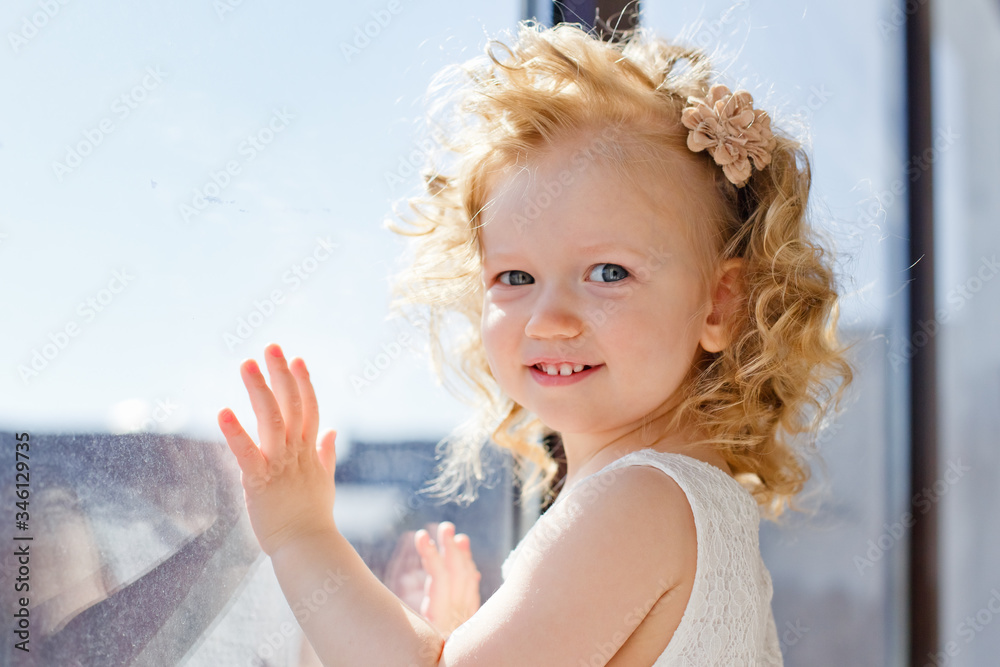 A beautiful little girl smiles and looks out the window. The child looks out the window. A portrait of a cheerful baby stands on the windowsill. A beautiful little girl smiles and looks out the window. The child looks out the window. A portrait of a cheerful baby stands on the windowsill.