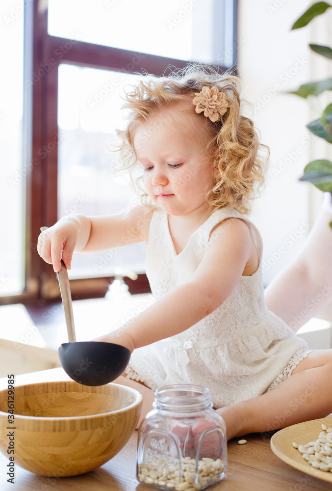 Obraz premium A little girl is sitting on a table in a bright Scandinavian-style kitchen. Playing with dishes, cereals, beans. The concept of children's development Montessori.