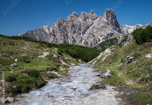River between meadow with mountain range in the background at Three Peaks nature reserve, blue sky during summer, South Tyrol Italy.