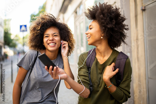two happy female black friends walking in city with mobile phone listening to music with earphones