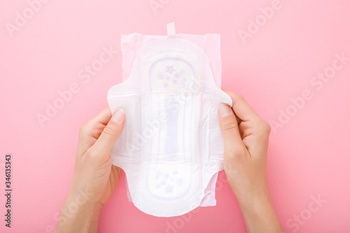Young woman hands holding opened white sanitary towel with wings on light pink table background. Pastel color. Closeup. Point of view shot. Top down view.