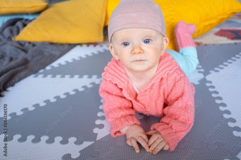 Adorable baby girl wearing pink jacket, crawling on floor, lying on belly with yellow cushions in background. Infant child in home interior. Childhood concept