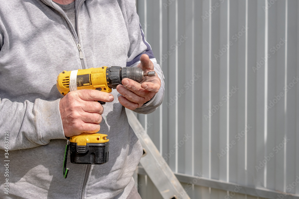 Yellow cordless screwdriver in corpus callosum male hands. Sunny day. The screwdriver is covered with scratches indicating its frequent use.