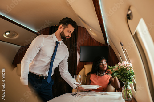 selective focus of handsome air steward serving table near elegant african american woman in plane