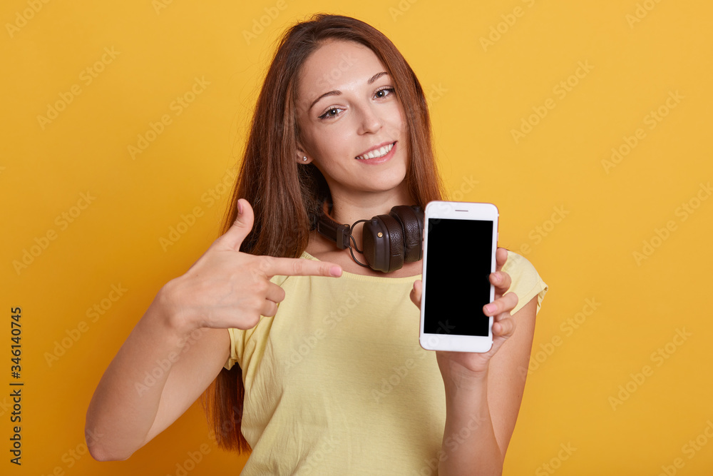 Horizontal shot of smiling woman pointing at blank device's screen holding in hands, lady with headphones around neck wearing casual t shirt, girl with long dark hair, against yellow studio wall. Horizontal shot of smiling woman pointing at blank device's screen holding in hands, lady with headphones around neck wearing casual t shirt, girl with long dark hair, against yellow studio wall.