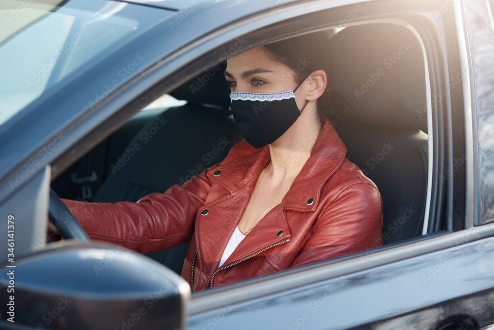 Naklejka premium Outdoor picture of confident busy young female driving car, wearing antibaterial mask, opening window, being in quarantine, having deals, spending time alone. People and coronavirus concept.