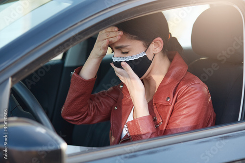 Picture of infected sick young female closing eyes, coughing, covering mouth, having fever, putting one hand on forehead, driving car, being ill with coronavirus. People and covid 19 concept. photo