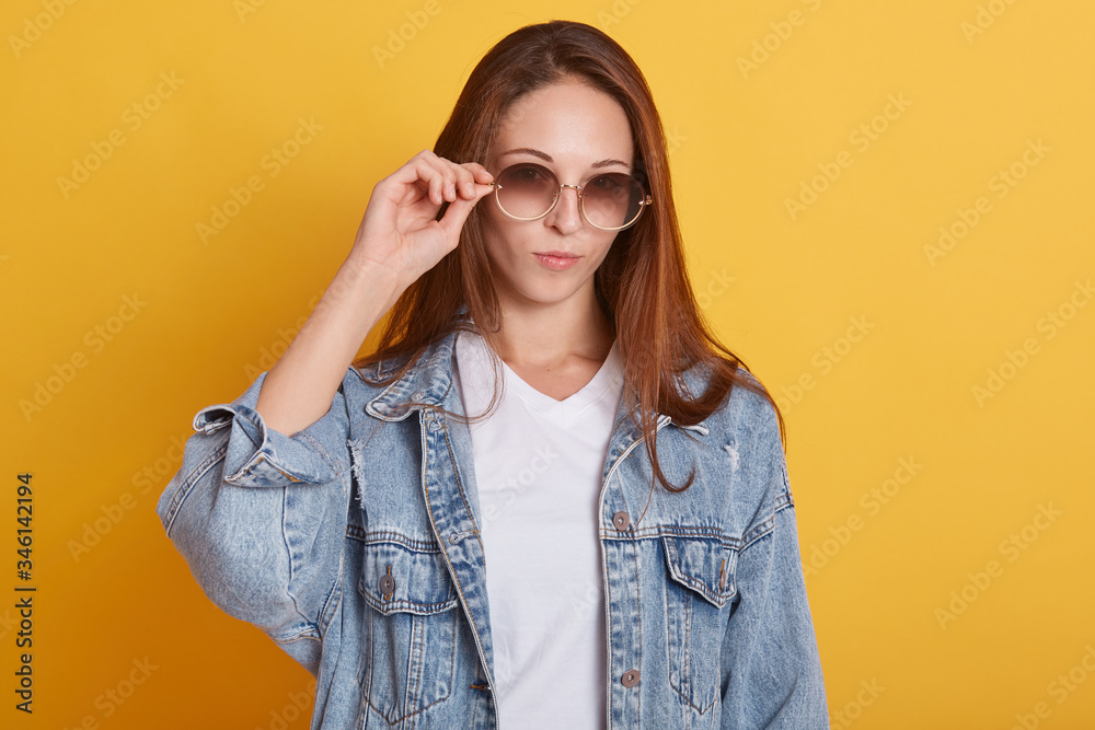 Close up portrait of glamorous stylish teenager posing isolated over yellow background in studio, wearing trendy sunglasses and denim jacket, being alone, having rest. Youth and style concept. Close up portrait of glamorous stylish teenager posing isolated over yellow background in studio, wearing trendy sunglasses and denim jacket, being alone, having rest. Youth and style concept.
