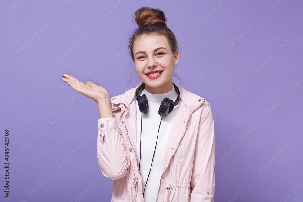 Horizontal shot of smiling adorable woman with hair bun standing with hands aside and with headset over neck, lady being in good mood, posing isolated over lilac studio background. Copy space. Horizontal shot of smiling adorable woman with hair bun standing with hands aside and with headset over neck, lady being in good mood, posing isolated over lilac studio background. Copy space.