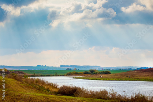 Autumn landscape with a river and clouds through which sunlight penetrates