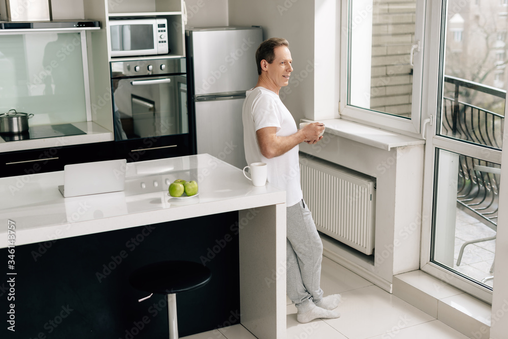 man holding bowl with cornflakes and looking at window near apples, laptop and cup on table man holding bowl with cornflakes and looking at window near apples, laptop and cup on table