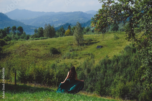 Happy, brunette girl running on the lawn. Carpathians mountains at the background. Young romantic woman in long, silk, green dress on the mountainside.