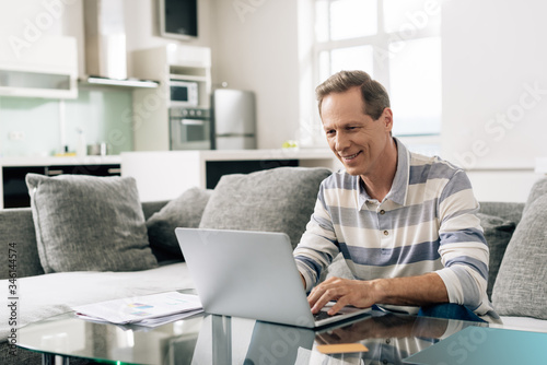 cheerful man smiling while using laptop near credit card in living room