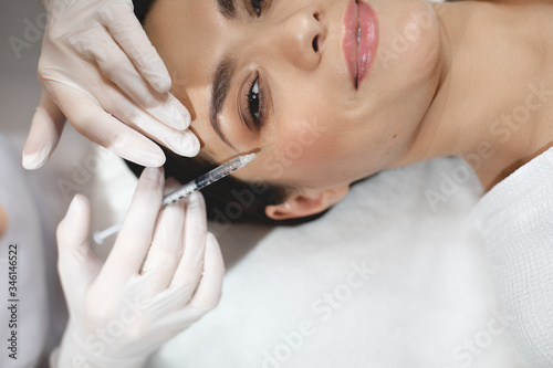 Cut view of happy positive young woman during botox injection process. Hands in white gloves holding syringe close to eye area and input some collagen under facial skin.