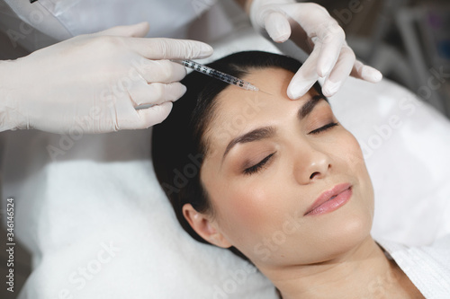 Young woman lying with closed eyes during botox injection process. Beautician in white gloves holding syringe at forehead area and input some collagen under skin.