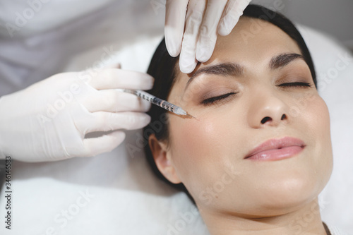 Young woman lying with closed eyes during botox or collagen injection at eye wrinkle area. Doing procedure accurate and careful.