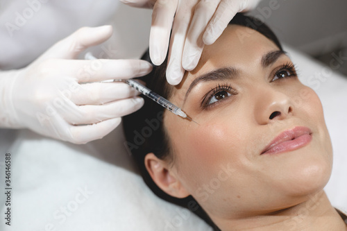 Process of injecting botox under facial skin at eye area with syringe. Young happy woman lying during procedure. Beautician does it carefully.