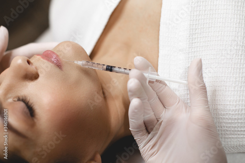 Side angle view of sleepy calm young woman during injection botox process. Hand in white glove inject some collagen from syringe.