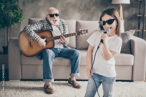 Photo of aged grandpa sitting comfy sofa playing guitar little pretty granddaughter hold mic singing acoustic song spend time together stay home quarantine modern living room indoors
