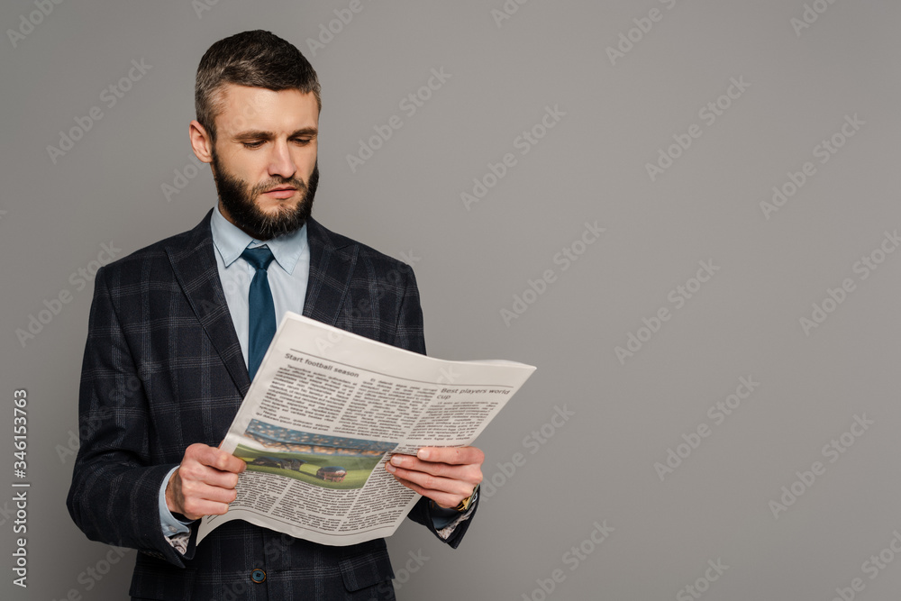 smiling handsome bearded businessman in suit with newspaper on grey background