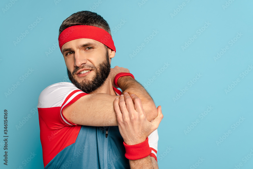 Naklejka premium bearded stylish sportsman warming up arm on blue background