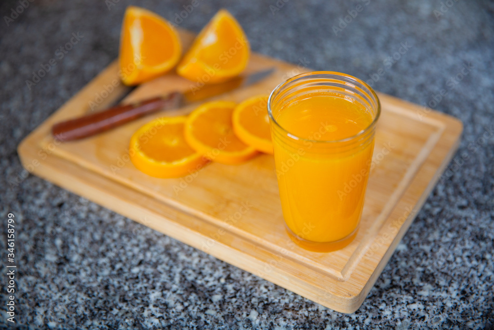 Fototapeta premium Glass of fresh orange juice standing on wooden board in focus and group of orange pieces cutting with knife out-of-focus. Close-up side view. Citrus fruit, vitamin and healthy food concept