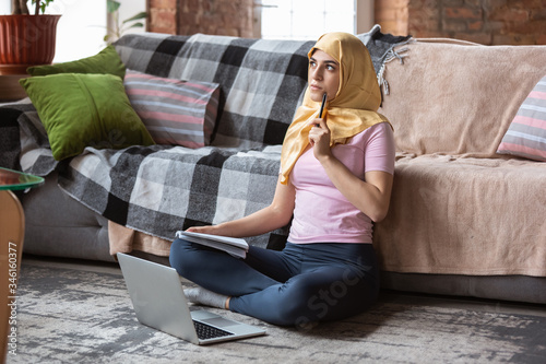 A pretty young muslim woman at home during quarantine and self-insulation. Arabian female model spending time useful being isolated. Concept of healthcare, communication, education during pandemic. photo