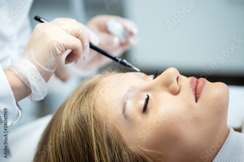 A young woman lies and master in a white uniform does eyebrow makeup in a beauty salon. The use of permanent makeup on brows. The wizard works with eyebrows. Semi-permanent makeup. Close up portrait